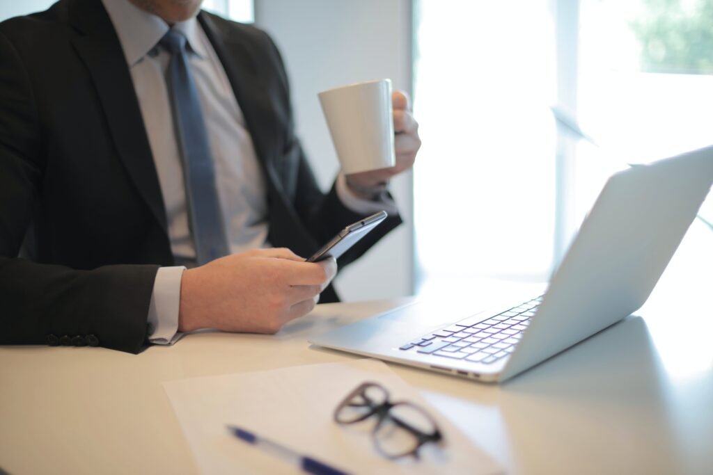 Hombre con traje de ejecutivo sentado frente a la laptop tomando taza de café y viendo el celular